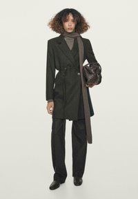 Mørkegrå skræddersyet frakke med bælte i taljen, kombineret med matchende bukser. En lysegrå rullekrave og brun flettet håndtaske fuldender looket.