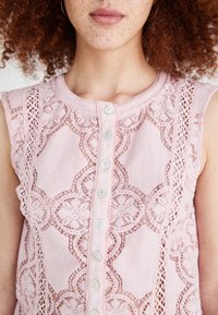 Blouse rose clair avec des panneaux en dentelle ornés de motifs floraux, fermeture à boutons et col rond. La texture du tissu est douce et lisse.