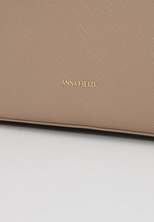 Osoba v tmavém kabátě nese velkou béžovou tašku s ramenními popruhy přes ruku na neutrálním pozadí.