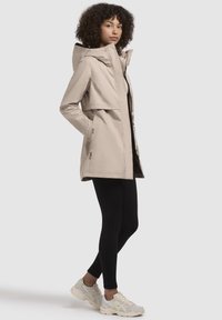 Beige lichtgewicht jas met capuchon, met een flap aan de voorkant, twee zijzakken, een gladde textuur en een strakke uitstraling, gedragen over zwarte leggings.
