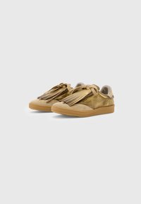 Un paio di sneakers metalliche oro con punte in suede beige, dettagli a nappina sopra i lacci e suole in gomma color marrone chiaro su uno sfondo bianco.