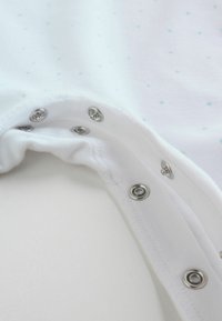 Indumento in cotone bianco con motivo di stelle azzurre, dotato di chiusure a pressione argento lungo il bordo inferiore e una texture morbida e liscia.