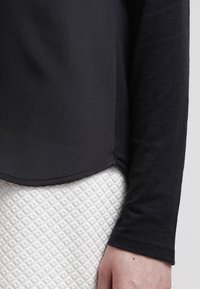Schwarzes Langarmoberteil mit glatter Textur und abgerundetem Saum, kombiniert mit einem weißen gemusterten Strickrock mit einem Diamantdesign.