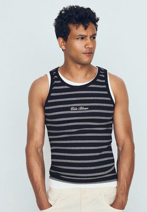 Jeune homme aux cheveux bouclés portant un débardeur rayé noir et blanc et un pantalon clair, regardant sur le côté avec les mains dans les poches.