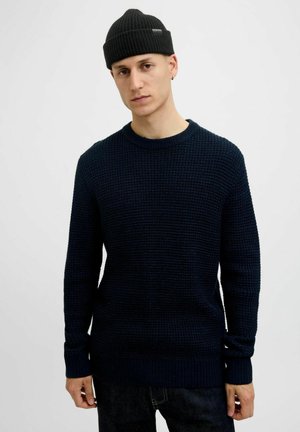 Navy knitted sweater with a textured weave, round neckline, and ribbed cuffs. Worn with dark jeans and a black ribbed beanie.