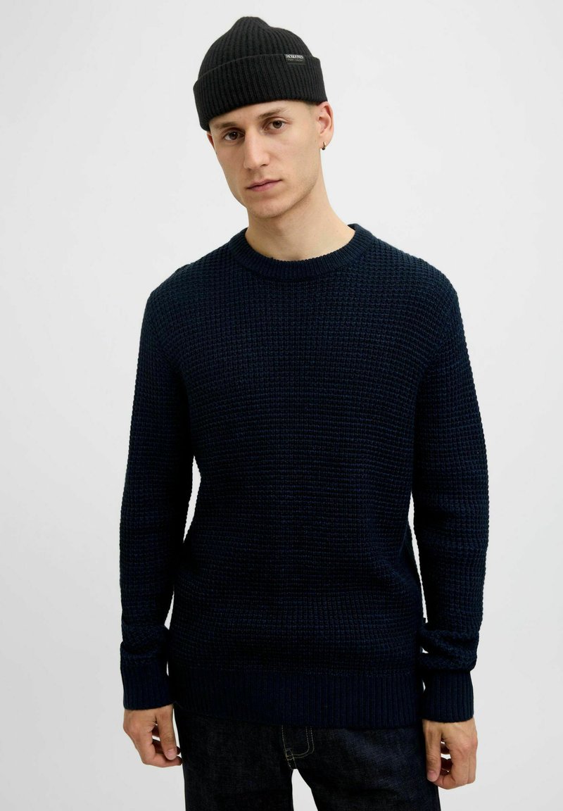 Pull en maille bleu marine avec un tissage texturé, col rond et poignets côtelés. Porte avec un jean foncé et un bonnet noir à côtes.