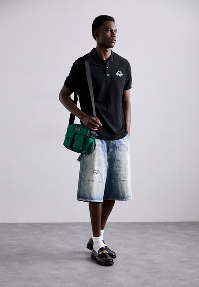 Svart polo skjorte, lyseblå denimshorts, hvite sokker, svarte sko. Grønn skulderveske med flere lommer. Nøytral bakgrunn.