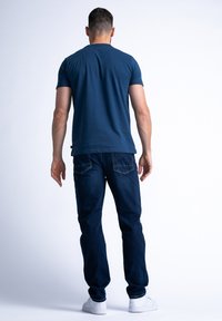Marineblå bomulds T-shirt med korte ærmer, parret med mørkeblå denim jeans. Modellen har hvide sneakers på og viser et afslappet look fra ryggen.