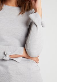 Jasnoszary dzianinowy sweter z okrągłym dekoltem, prążkowaną teksturą i kokardkami przy mankietach, zapewniający dopasowaną sylwetkę.