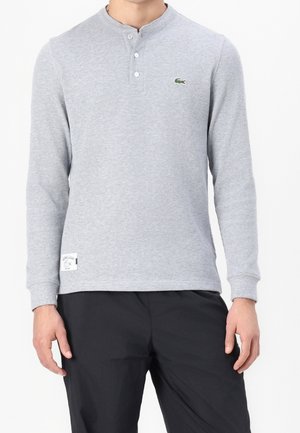 Mand iført lys grå langærmet henley-shirt med knapper og sorte bukser, med lille grønt krokodillelogo på brystet.