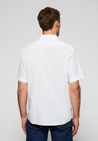 Camicia bianca a maniche corte realizzata in tessuto leggero. Presenta un colletto classico e un design pulito e semplice con orlo leggermente arrotondato.