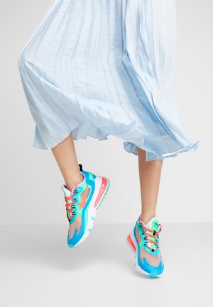 Jambes portant une jupe plissée bleu clair fluide et des baskets colorées turquoise, orange et rose, montrées en plein pas sur un fond uni.