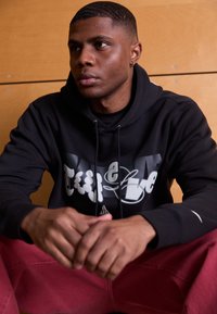 Felpa nera con stampa grafica che presenta lettere bianche e grigie. Il modello indossa pantaloni rossi, è seduto con le mani incrociate, su uno sfondo in legno.