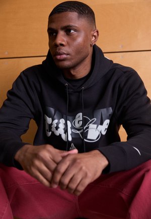 Felpa nera con stampa grafica che presenta lettere bianche e grigie. Il modello indossa pantaloni rossi, è seduto con le mani incrociate, su uno sfondo in legno.