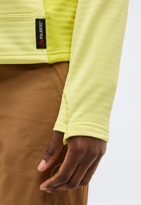 Makro posnetek temnopolte roke blizu manšete rumene majice z dolgimi rokavi z oznako Polartec, ki počiva ob rjavih hlačah.