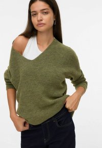 Vrouw in een olijfgroene off-shoulder trui over een witte tanktop, met donkere jeans, lange bruine haren en zilveren oorringen.