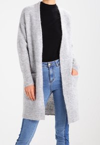 Ljust grå cardigan i ett mjukt, texturerat tyg, med öppen framsida, långa ärmar och två framfickor.