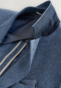 Modrý vzorovaný blazer s vyvýšeným límcem, který odhaluje tmavě modrou podšívku a béžový lem podél vnitřního švu.