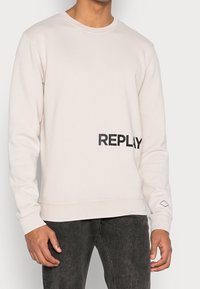 Ljust beige sweatshirt med rund halsringning, med en stor svart "REPLAY"-tryck på framsidan och en diamantlogotyp på ärmen.