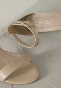 Sandales de couleur crème avec une semelle intérieure texturée, une lanière lisse et une bride de cheville réglable dotée d'une boucle dorée.