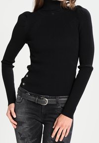 Pull noir côtelé à col roulé avec manches longues, agrémenté de boutons sur le côté et d'une coupe ajustée. Associé à un jean noir déchiré.