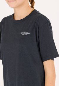 Tummansininen puuvillapaita, jossa on pyöreä pääntie ja lyhyet hihat. Paitassa on pieni valkoinen "Sports Club Les Arcs" -logo rinnassa.