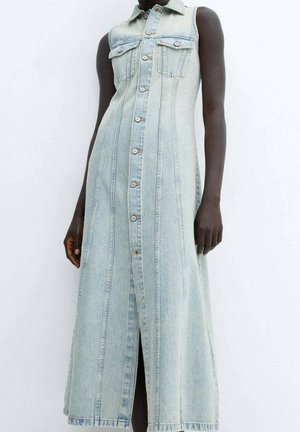 Robe maxi en denim bleu clair sans manches avec boutons à l'avant, poches poitrine et coutures structurées portée par une personne debout sur un fond uni.