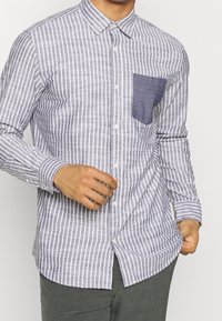 Randig skjorta i grått och vitt, med en ficka i blå denim. Lång ärm, krage och en slim fit.