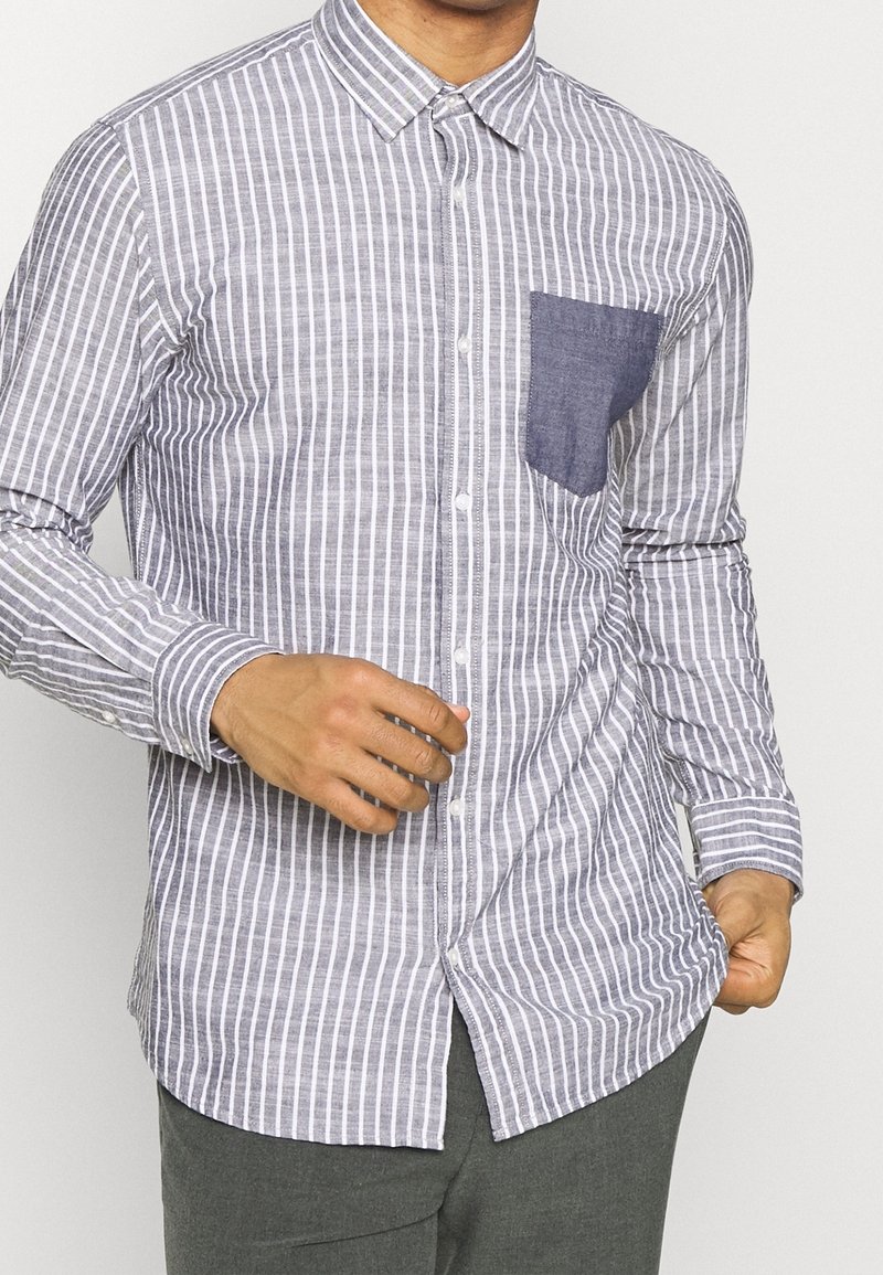 Randig skjorta i grått och vitt, med en ficka i blå denim. Lång ärm, krage och en slim fit.