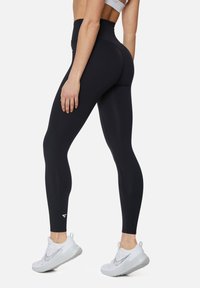 Hoge taille zwarte legging van rekbaar materiaal, met een strakke pasvorm en een klein wit logo bij de enkel, gecombineerd met witte sneakers.