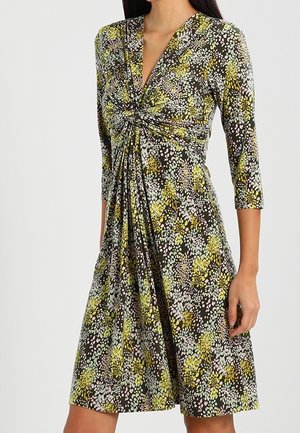 Femme portant une robe longue jusqu'aux genoux, à manches longues, avec un nœud torsadé à la taille et un motif floral abstrait multicolore.
