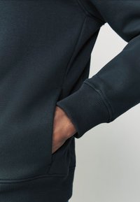 Tmavomodrá mikina s bočným vreckom, rebrovanými manžetami a mäkkým, textúrovaným povrchom. Zakrivené švy zlepšujú dizajn.