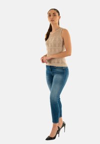 Top sin mangas de punto beige con un patrón texturizado, combinado con jeans ajustados azules y zapatos de tacón altos negros.