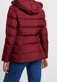 Person som bär en vinröd vadderad jacka med huva, kombinerad med blå jeans, sedd bakifrån mot en enkel ljus bakgrund.