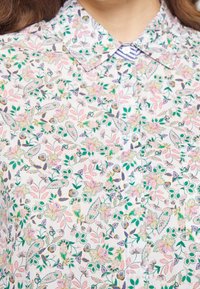 Chemise en tissu blanc avec un motif floral en rose, vert et violet, dotée d'un col boutonné et d'un détail rayé subtil.