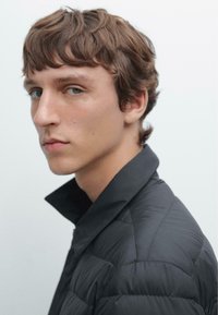 Schwarze Pufferjacke mit hohem Kragen, mit gestepptem Design und eleganter Optik. Das Model hat kurzes, zerzaustes braunes Haar und helle Augen.