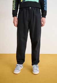 Black, relaxed-fit pants with an elastic waistband and side pockets, paired with light blue sneakers on a yellow backdrop.