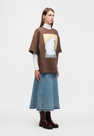 Bruine oversized T-shirt met een witte kattenafbeelding, gecombineerd met een lichtblauwe denim rok met een gebogen naad, en glanzende bordeauxrode enkellaarzen.