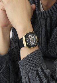 Reloj de pulsera negro y dorado con caja cuadrada, correa de goma texturizada negra, tres subesferas y marcaciones blancas, combinado con un suéter de punto gris.