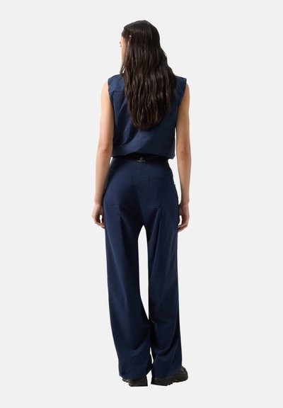 Frau mit langem dunklem Haar, die einen ärmellosen marineblauen Oberteil und weit geschnittene marineblaue Hosen trägt, steht mit dem Rücken zu einem schlichten Hintergrund.