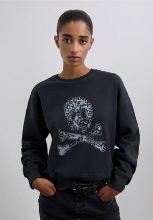 Un sweat à capuche noir affichant un graphique de crâne et d'os croisés avec un détail texturé. Le design intègre divers motifs avec une finition brillante.