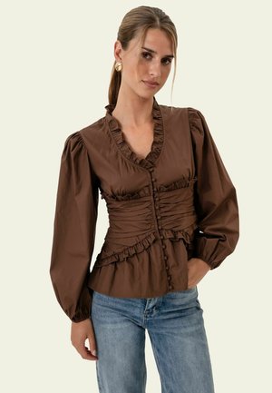 Frau mit hellbraunen, zurückgebundenen Haaren trägt eine braune, langärmelige Bluse mit Rüschen und blaue Jeans, steht mit einer Hand in der Tasche.