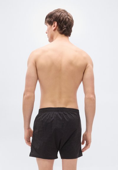 Μαύρα μαγιό σορτς με υφή, ελαστική μέση και κοντό μήκος, εμφανιζόμενα από πίσω σε μοντέλο με ουδέτερο φόντο.