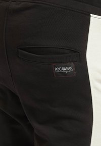 Juodos sportinės kelnės su baltu šoniniu paneliu, turinčios nugarinę kišenę ir raudonai akcentuotą etiketę su užrašu "ROCAWEAR." Minkšta, tekstūruota medžiaga.