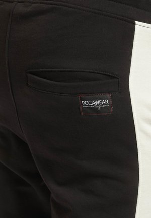 Sorte sweatpants med en hvid sidepanel, der har en baglomme og et label med røde detaljer, som viser "ROCAWEAR." Blødt, tekstureret stof.
