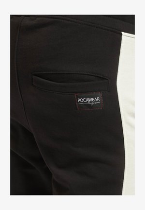Sorte sweatpants med en hvid sidepanel, der har en baglomme og et label med røde detaljer, som viser "ROCAWEAR." Blødt, tekstureret stof.