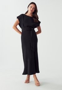Musta, hihaton midi-mekko, jossa on leveä pyöreä kaula-aukko, vyötärönauha ja virtaava helma. Sileä pinta, pehmeä laskeutuminen ja minimalistinen muotoilu.