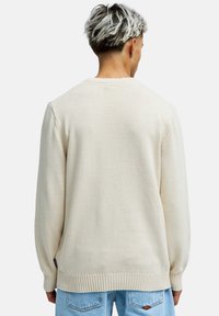 Mies, jolla on lyhyet, värjätyt hiukset, yllään kermaisen värinen neulepaita ja vaaleansiniset farkut, katsomassa poispäin yksinkertaista valkoista taustaa vasten.