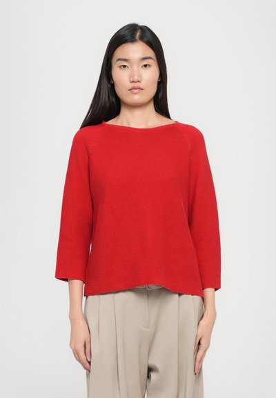 Pull en tricot rouge avec un décolleté large et des manches trois-quarts, présentant une coupe décontractée et une surface texturée. Porté avec un pantalon beige.