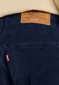 Pantaloni in velluto a coste blu navy con texture a coste visibili, dotati di un'etichetta in pelle marrone con dettagli del marchio e un piccolo logo rosso sulla tasca sinistra.